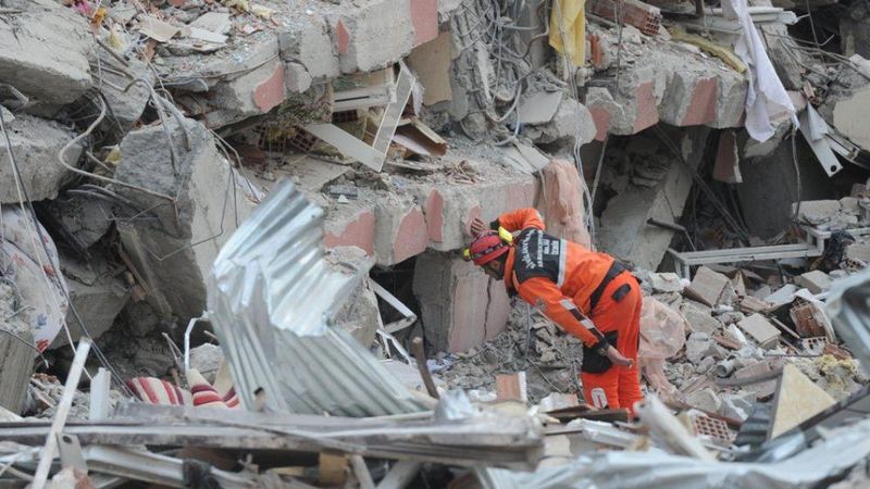 A rescue worker in an orange jumpsuit searching through earthquake rubble